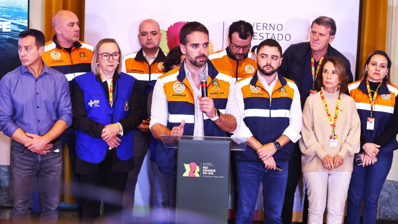 Leite ao microfone, falando e gesticulando atras de um púlpito em coletiva sobre enchentes de 10 de maio de 2024. Atrás deles estão diversos secretários de Estado.