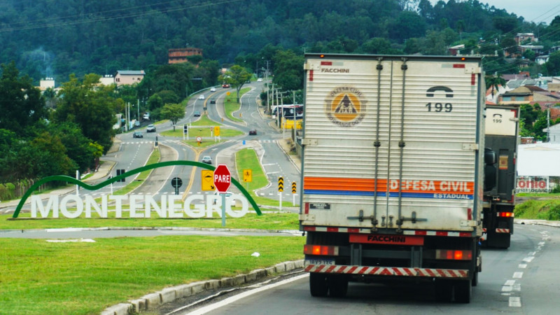 Caminhões da Defesa Civil Estadual visto de trás na estrada em local onde se vê, mais às esquerda, no centro de uma rótula, um letreiro com o nome da cidade: Montenegro