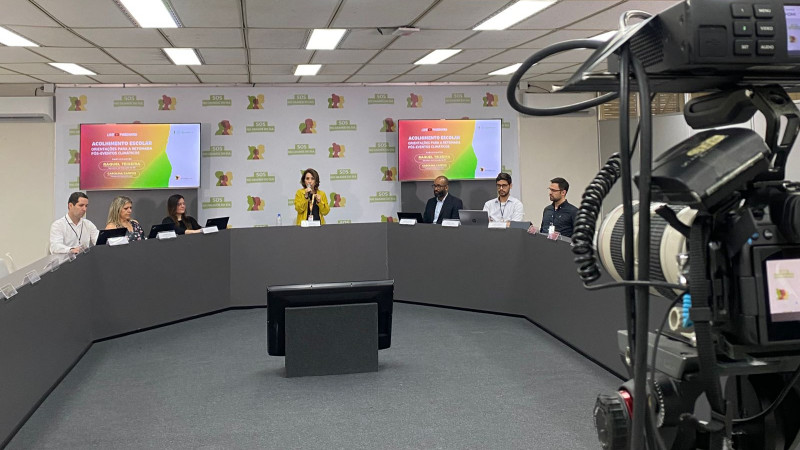Live sobre acolhimento escolar e orientações da Seduc após enchentes de 2024. Raquel e outras pessoas durante a transmissão. No canto da imagem uma câmera de vídeo capta imagens que são transmitidas.
