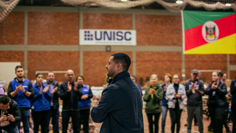No Ginásio da Universidade de Santa Cruz do Sul (Unisc), um homem aparece de costas, falando para um grupo de pessoas o aplaudem.