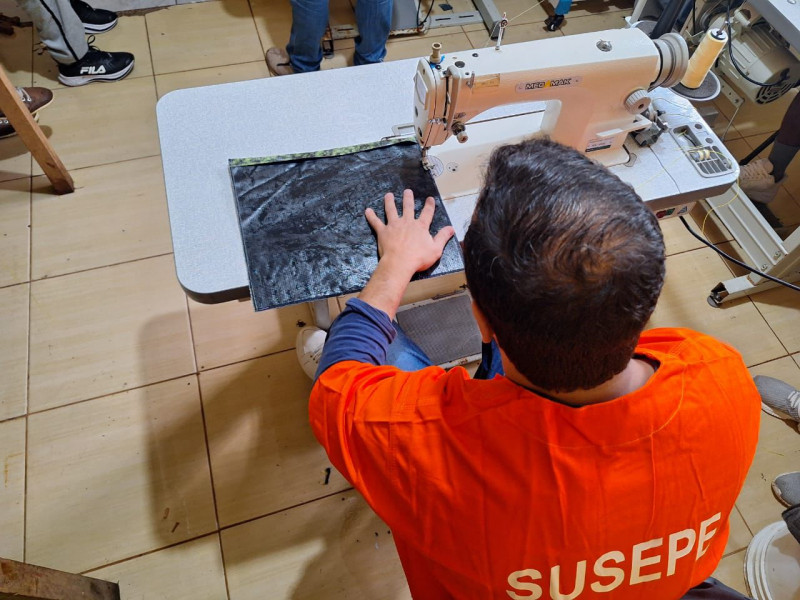 Um apenado de costas utilizando a máquina de costuras confecciona uma bolsa para kit de maternidade.