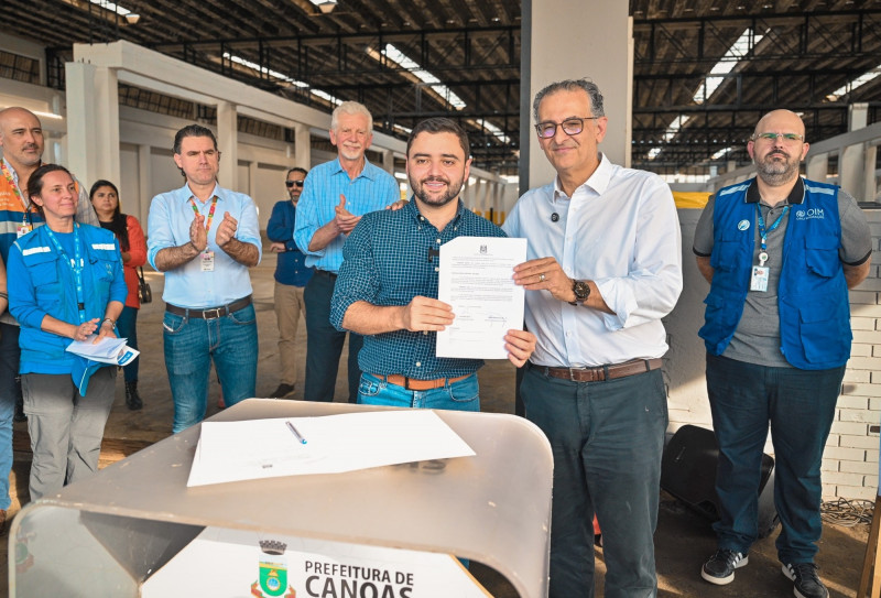 Formalização dos Chás em Canoas. Gabriel e o prefeito seguram o documento assinado em foto posada. Há pessoas atrás deles.
