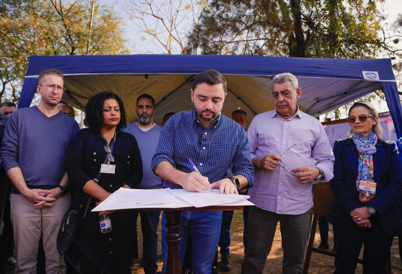 Formalização dos CHAs em Porto Alegre 2024. Gabriel assina o documento sendo observado por pessoas atrás dele.
