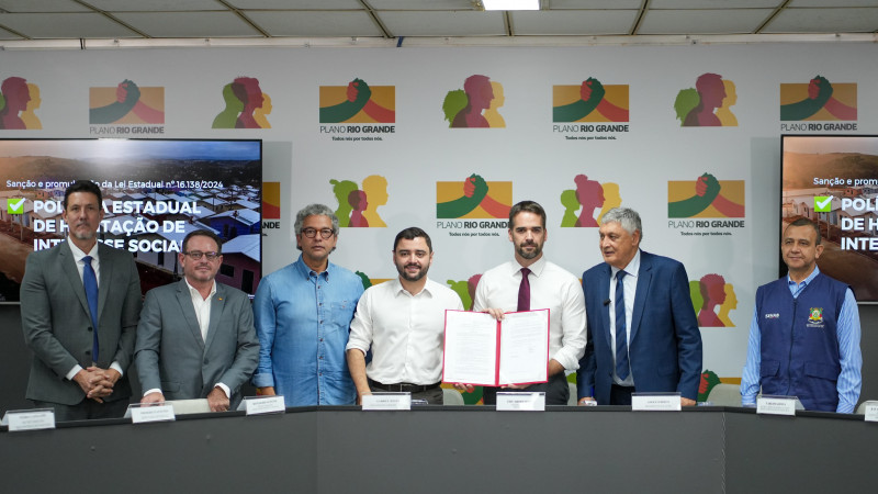 Foto posada. Leite e Gabriel estão no centro de pé, ao lado de outros cinco representantes do Estado e políticos. Leite segura o documento assinado, mostrando-o para a câmera.