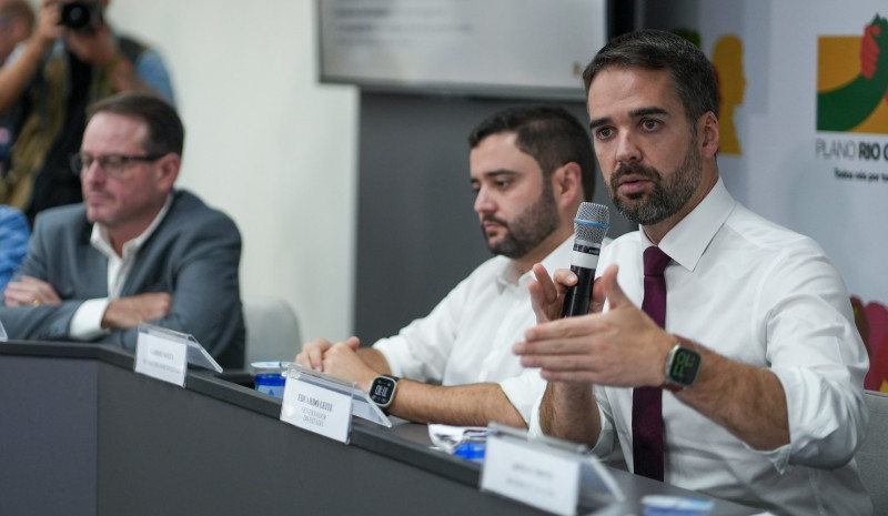 Leite aparece sentado, falando ao microfone e gesticulando com a mão, num ângulo que o pega meio de lado. Ao fundo está Gabriel, também sentado.