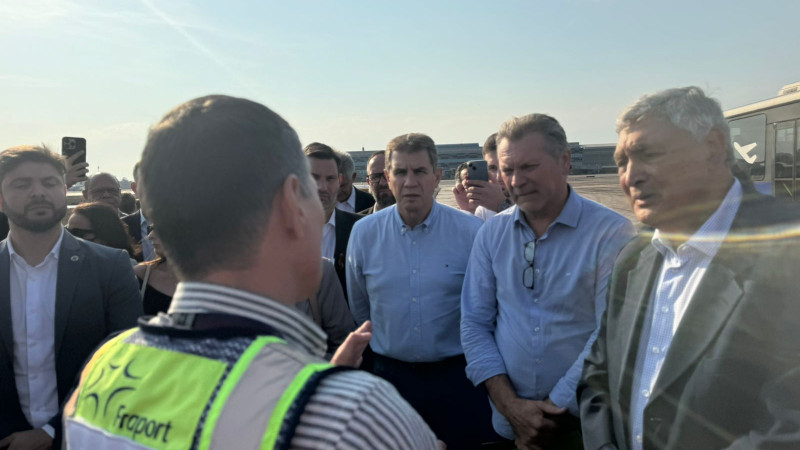 Foto em ambiente aberto, na pista do aeroporto. Um grupo de cinco homens é enquadrado, de pé, a partir da cintura, de frente para a câmera. Eles conversam com outro homem, que aparece de costas, com um colete verde da Fraport.