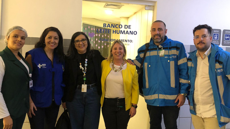 Foto posada em ambiente hospitalar. Quatro mulheres e dois homens estão de pé, enquadrados em plano americano, sorrindo para a câmera. Atrás, está uma porta de vidro onde está escrito Banco de Leite Humano.