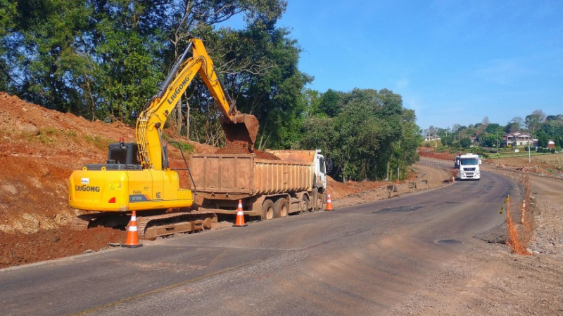Foto em ambiente aberto de uma escavadeira jogando terra na caçamba de um caminhão, ambos no acostamento de uma estrada, junto a um aterro de terra. Ao fundo, aparecem árvores.