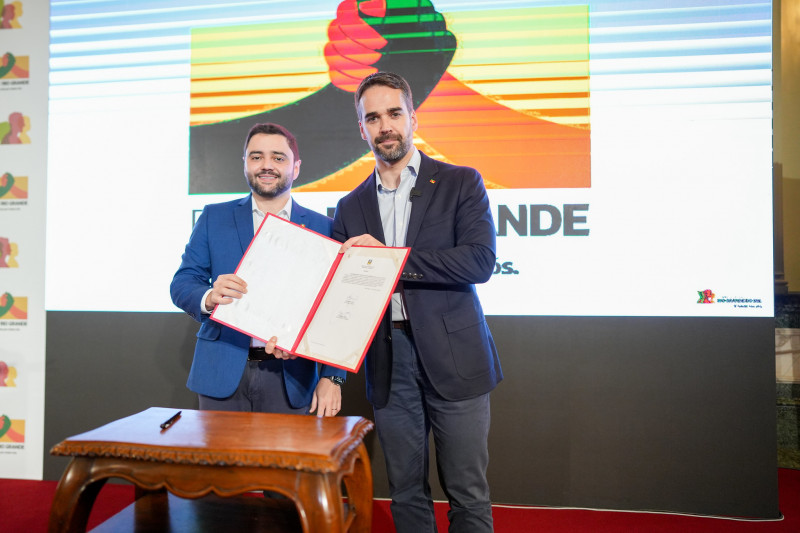 Gabriel e Leite posam para a câmera, de pé, exibindo o documento assinado e sorrindo. Ao fundo, a logo do Plano Rio Grande.
