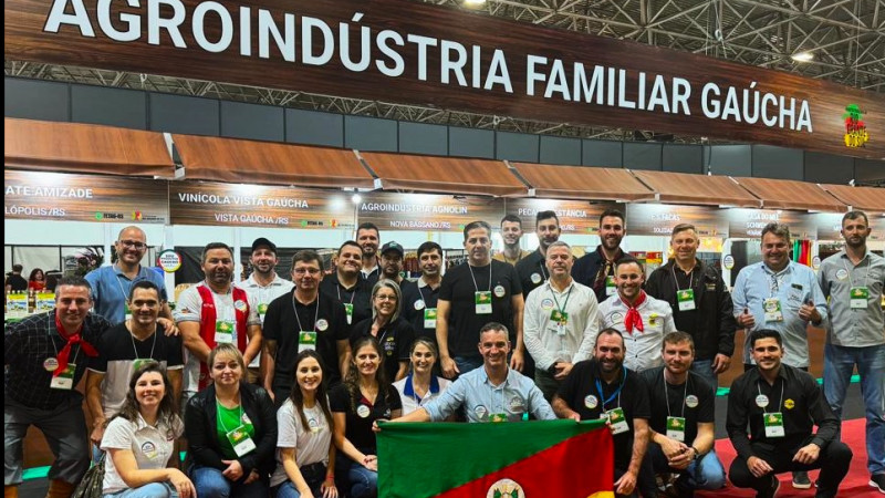 Foto posada com os produtores que foram à feira na frente do pavilhão da Agroindústria Familiar Gaúcha na Expotchê 2024. Um deles segura uma bandeira do Rio Grande do Sul.