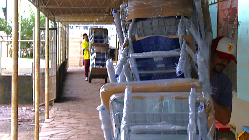 Entrega de mobiliário na Escola Guarani. Dois homens em movimento em um dos corredores externos da escola carregando em carrinhos diversas mobílias para dentro da escola.