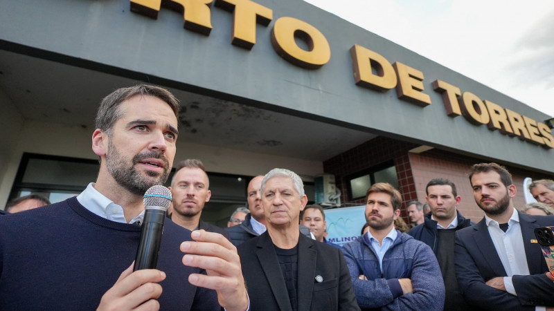 Governador falando ao microfone rodeado de muitas pessoas em área externa na entrada do Aeroporto de Torres.