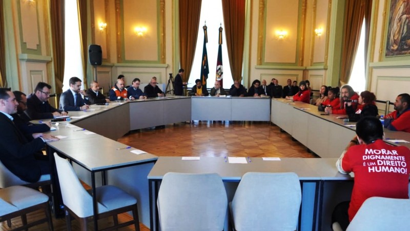 Reunião com Movimento de Luta nos Bairros, Vilas e Favelas. Várias pessoas reunidas em torno de uma grande mesa em formato de U.