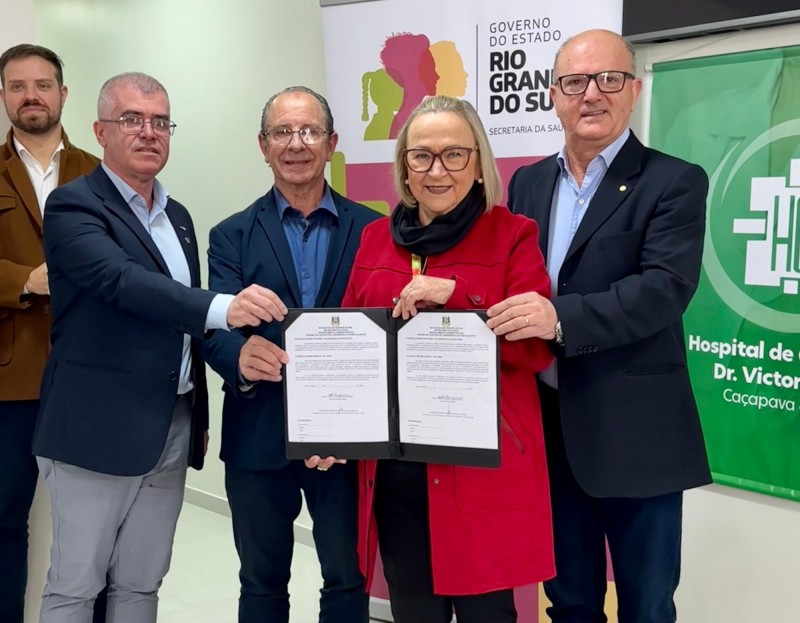 A imagem mostra cinco pessoas posando para uma foto, sendo uma delas a secretária segurando o documento assinado. Ao fundo, há banners do governo do Estado do Rio Grande do Sul, especificamente da Secretaria da Saúde, e do Hospital de Caridade Dr. Victor Lang de Caçapava do Sul. Todos estão vestidos formalmente.