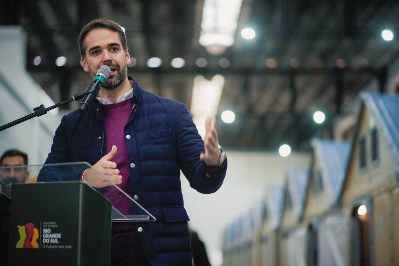 Leite fala ao microfone em um pódio com o logotipo do Governo do Estado do Rio Grande do Sul. Ele veste um casaco azul e um suéter roxo. O fundo mostra um ambiente interno com luzes e as estruturas das casas.