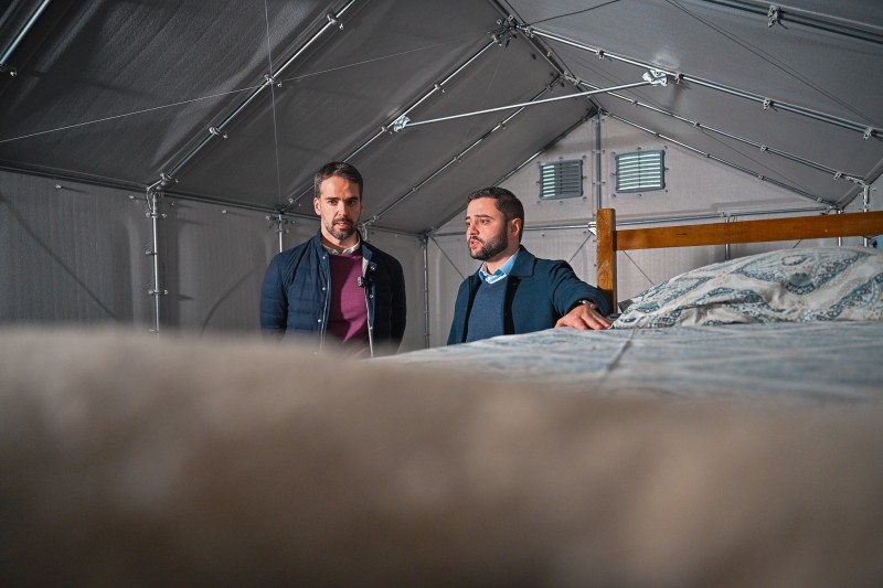 Leite e Gabriel estão em pé dentro de uma tenda, olhando para uma cama. Ambos vestem casacos escuros e estão em uma acomodação temporária.