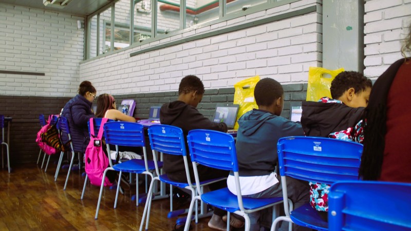 Cinco crianças aparecem de costas, sentadas em carteiras dispostas junto à parede do interior da sala de um abrigo. Elas estão com notebooks e parecem escrever em cadernos.