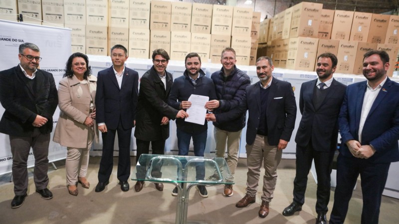 Entrega de kits de geladeiras e fogões. Foto posada. Ao centro, Gabriel Souza segura o documento junto com Beto Fantinel e o executivo da empresa. Há outras pessoas perfiladas na foto à direita e à esquerda.