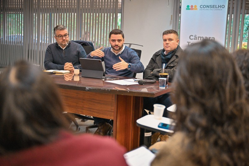 Gabriel aparece sentado junto a uma mesa, entre dois homens sentados, gesticulando enquanto fala para a audiência. No primeiro plano, aparecem duas mulheres de costas, sentadas, acompanhando a fala.