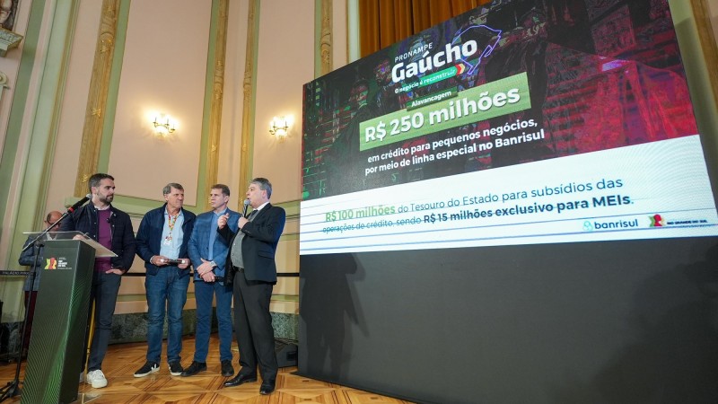 No lançamento do Pronampe Gaúcho, mais à esquerda da imagem, Leite atrás de um púlpito olha um homem falando em pé ao microfone. Três outros homens aparecem na foto. Mais à direita, uma tela exibe o nome e o valor do programa.