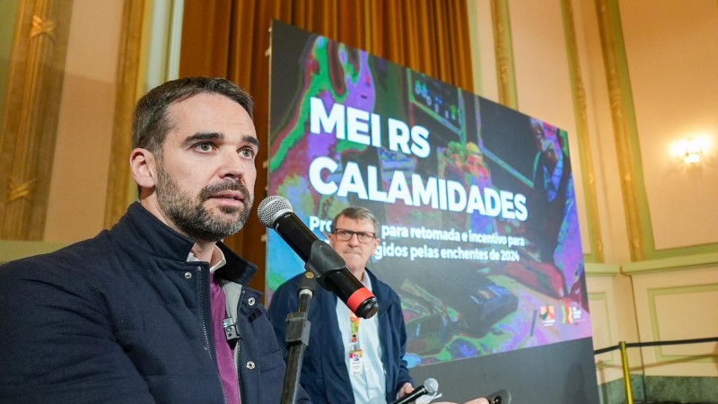Mais à esquerda Leite fala, em close, ao Microfone. Um homem o observa e mais à direita aparece o nome da iniciativa em uma tela grande: MEI RS Calamidades 2024