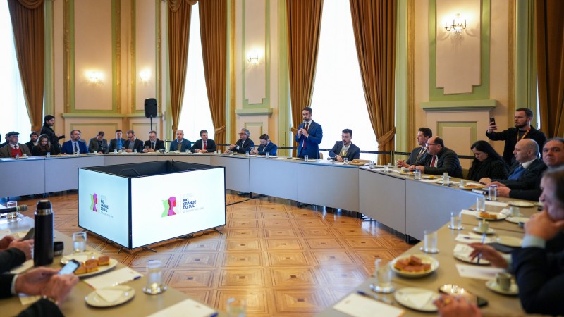 A foto mostra a reunião em uma sala ampla e bem decorada no Palácio Piratini, com cortinas douradas e painéis nas paredes. As pessoas estão sentadas ao redor de uma mesa em formato de "U", com o centro ocupado por um painel que exibe a logo do governo. Leite está de pé, falando ao microfone, enquanto os outros participantes estão sentados, escutando e tomando notas.
