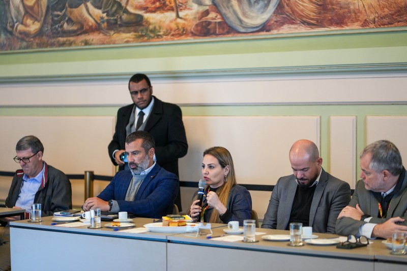 A foto mostra uma reunião formal com várias pessoas sentadas ao redor de uma mesa. Danielle fala ao microfone. A mesa tem copos, xícaras e pratos com comida. Ao fundo, um homem de pé observa a reunião. A sala é decorada com uma grande pintura na parede.