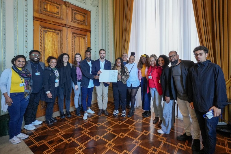 Foto posada, Leite, rodeado de diversas pessoas, está em pé e juntamente com duas outras pessoas segura o documento recé-assinado.
