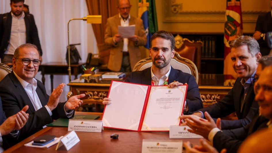 Leite segura o documento recém assinado e é aplaudio pelas outras pessoas que estão junto com ele sentados à mesa de reunião.
