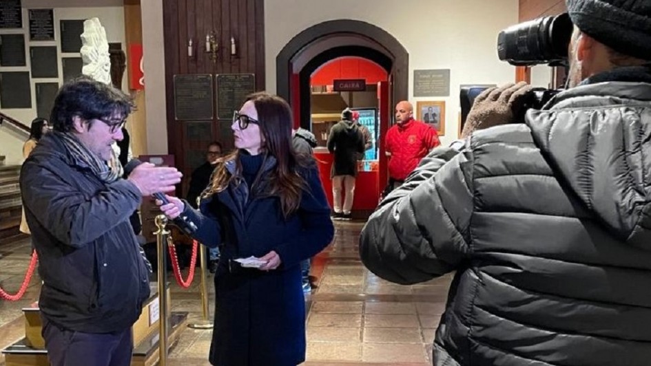 Imagem de cobertura anterior da TVE do Festival de Cinema de Gramado. À esquerda uma mulher entrevista um homem segurando um microfone que aponta para a boca dele. À direita, está o cinegrafista de costas segurando a câmera enquanto registra a entrevista.