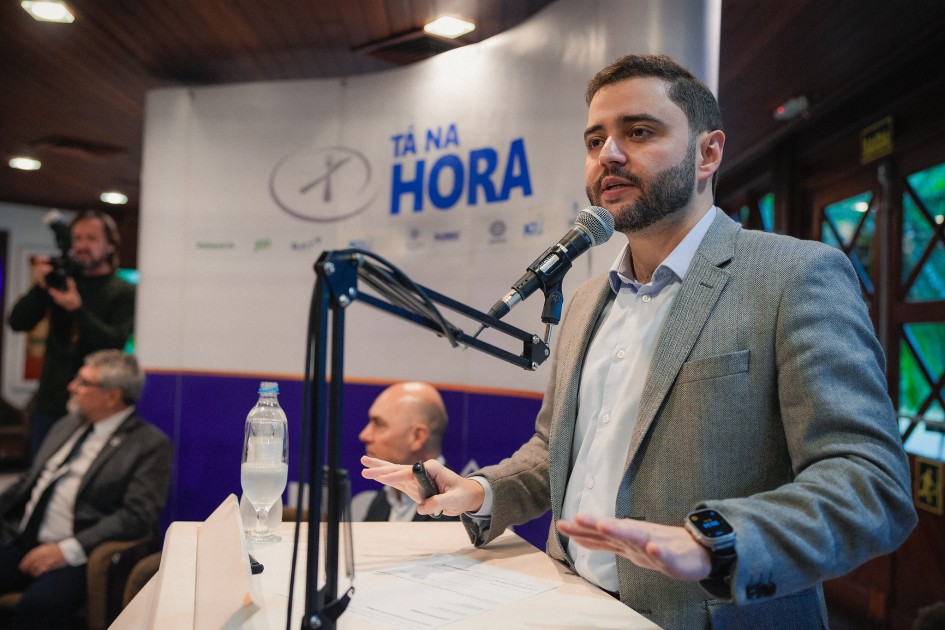 A imagem mostra Gabriel falando em um púlpito, com um microfone diante de si. Ele gesticula com as mãos enquanto fala, e ao fundo há um banner com o texto "Tá na Hora". No plano de fundo, duas outras pessoas estão sentadas.