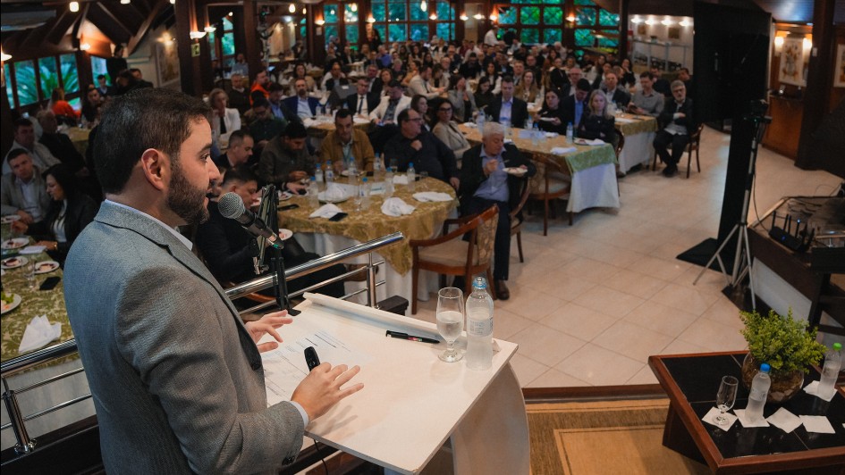 A imagem mostra Gabriel falando em um púlpito com um microfone. Ele está se dirigindo a uma grande audiência sentada em mesas redondas, em um salão fechado bem iluminado. As mesas estão cobertas com toalhas verdes, e há várias pessoas na sala, incluindo homens e mulheres.