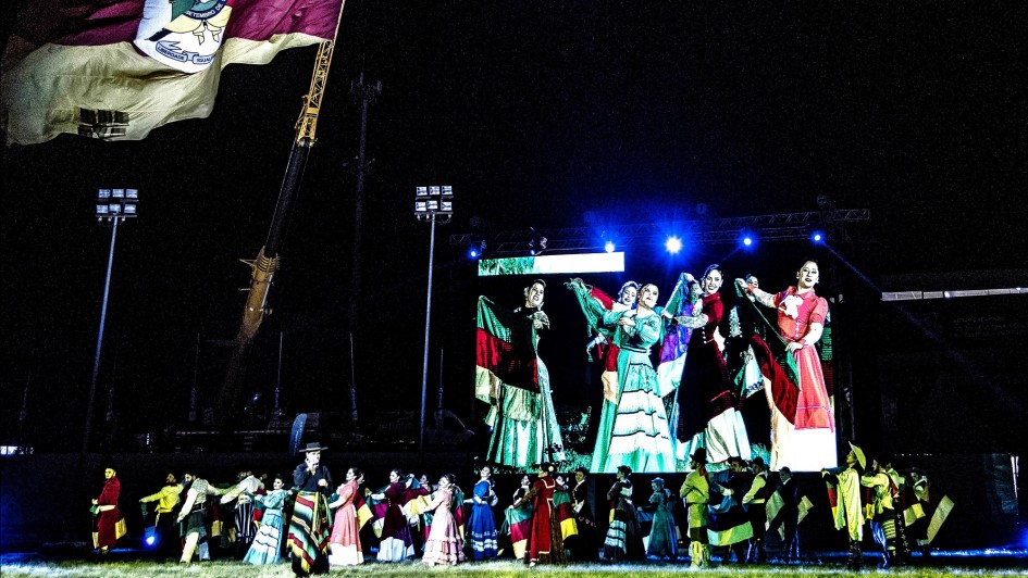 Imagem de uma apresentação anterior da Ópera Gaúcha na Expointer, com diversas pessoas dançando e um enorme telão atrás mostrando detalhes da dança.