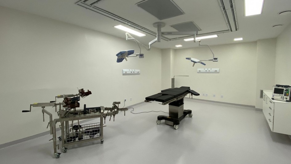 A imagem mostra uma sala cirúrgica moderna, equipada com mesas cirúrgicas, iluminação suspensa, e equipamentos médicos. A sala é ampla, com paredes e piso claros, e possui um ambiente bem iluminado, adequado para procedimentos cirúrgicos. No canto direito, há um monitor ou desfibrilador, enquanto à esquerda, há uma mesa cirúrgica ajustável com vários suportes e alavancas.