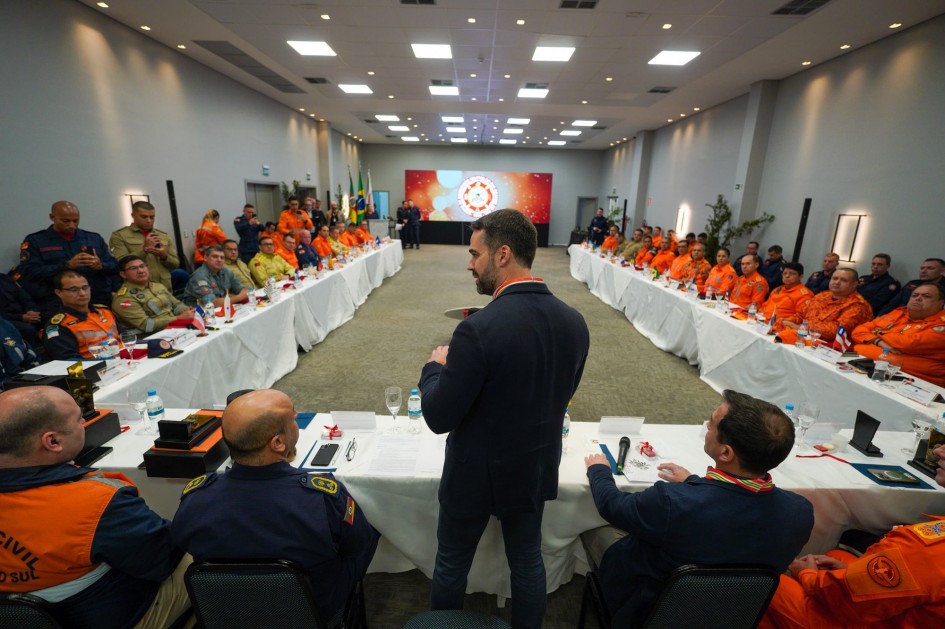 Leite ao microfone e levemente de costas falando para uma plateia de bombeiros militares disposto ao seu lado e às sua frente sentados em uma enorme mesa em formato de U.
