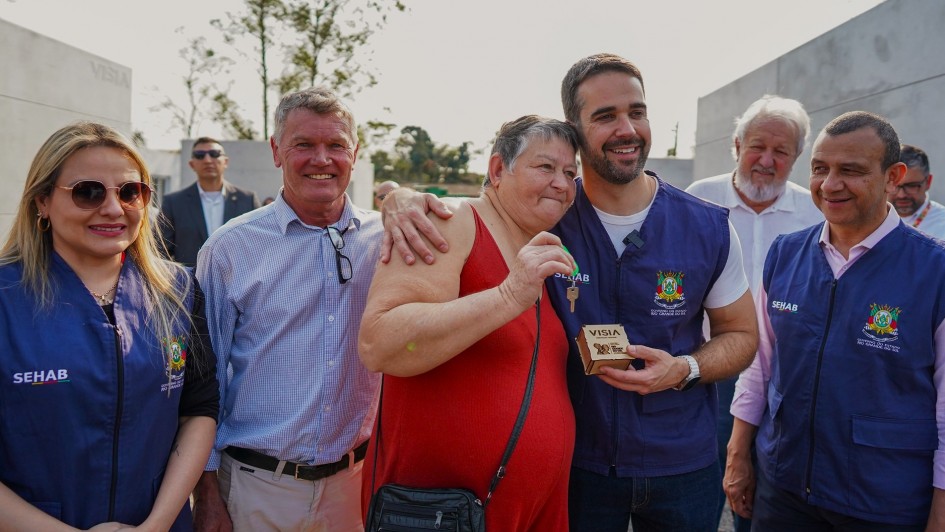 Leite entregando as chaves para uma mulher. Ele a abraça e há outras pessoas participando da foto e observando a entrega.
