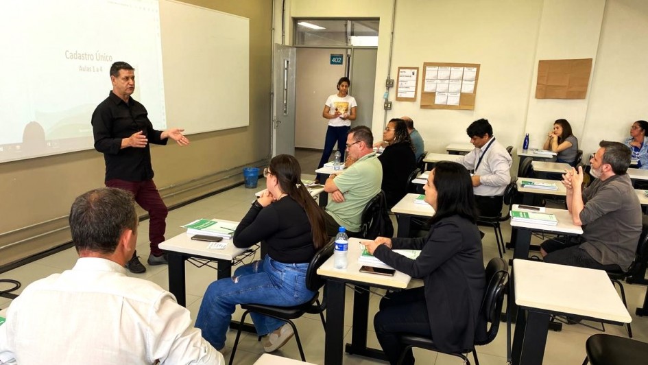 Professor dando aulas com seus alunos de perfil e sentados em cadeiras prestando atenção ao que ele fala.