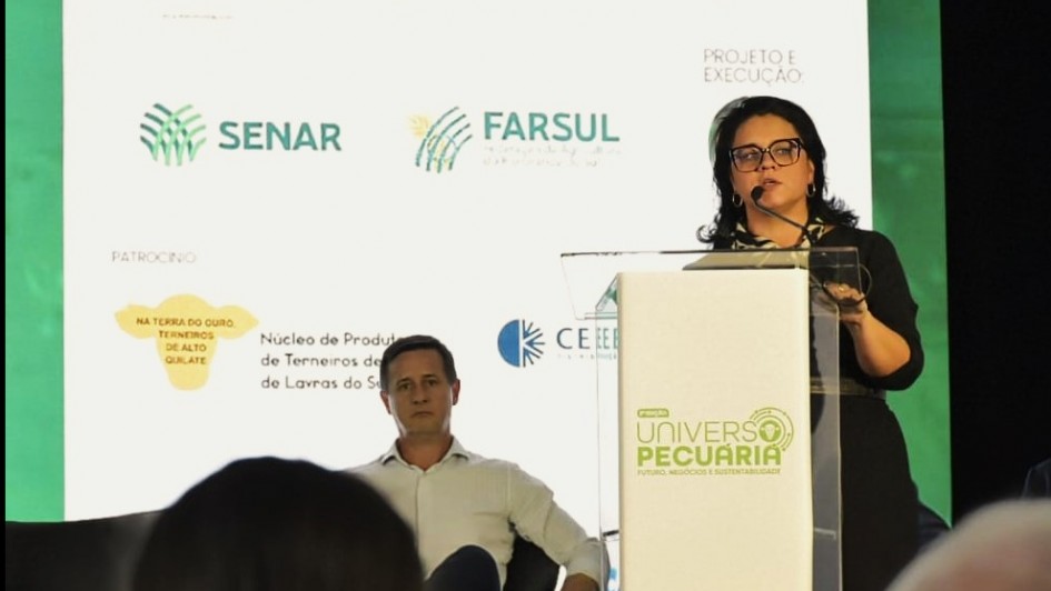 Marjorie falando ao microfone atrás de um púlpito e um pouco mais atrás Kuhn está sentando observando.