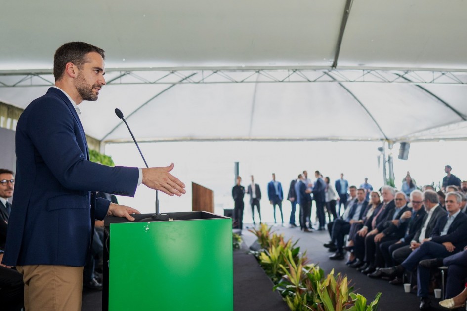 Leite de perfil ao microfone falando atrás de um púlpito para a plateia, que está mais à direita da imagem.