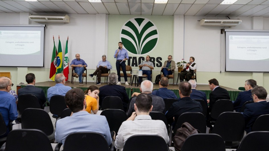 Leite falando ao microfone durante reunião com a Fetag-RS. Há cinco homens sentados ao lado de Leite em cima do palco e parte da plateia pode ser vista de costas.