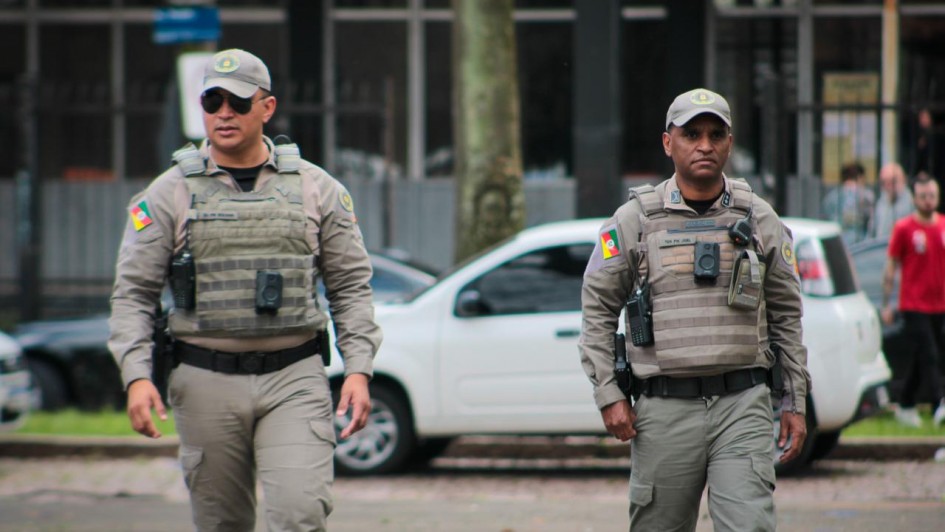 Dois polícias, em plano americano e de frente, caminhando na rua. Eles estão sérios e uniformizados, usando coletes com as câmeras.