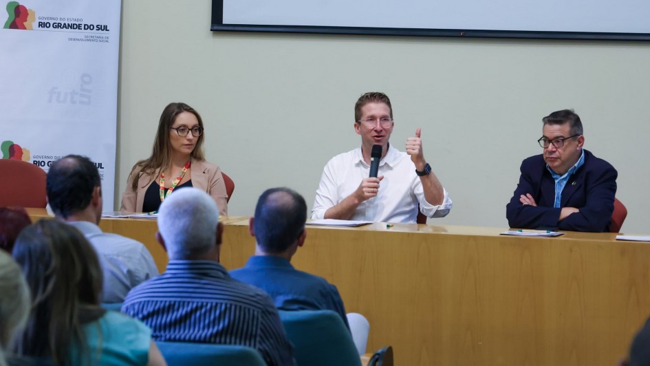 Imagem de três pessoas atrás de uma bancada. Uma delas fala ao microfone. Parte da plateia é vista de costas no workshop da Consulta Popular da Sedes.