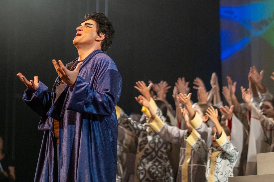 Foto lateral dos artistas se apresentando no palco. À esquerda, um homem de trajes azuis e maquiagem canta de forma dramática, com as mãos erguidas na altura do peito. Ao fundo, crianças em trajes claros também cantam, com as mãos erguidas para cima.
