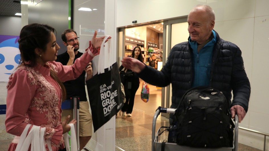 Senhor de casaco recebe ecobag de mulher vestida com trajes gaúchos no saguão do aeroporto. Ele empurra um carrinho com malas e sorri.