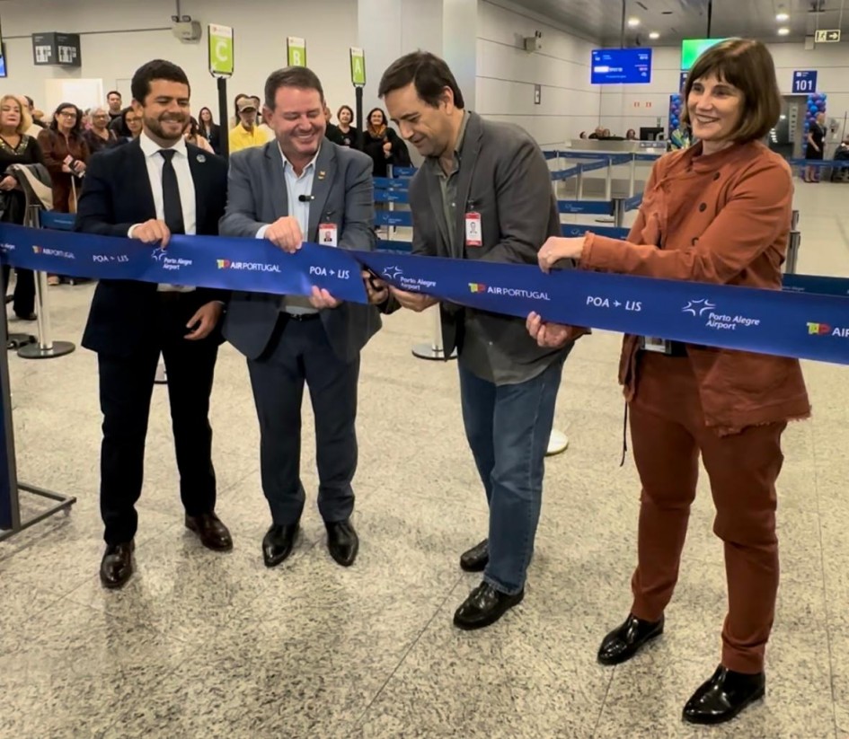 Quatro pessoas, dentre elas o secretário, cortam uma faixa simbólica no saguão do aeroporto, em alusão à retomada. A faixa é de cor azul, e nela está escrita Tap Air Portugual e POA + LIS.