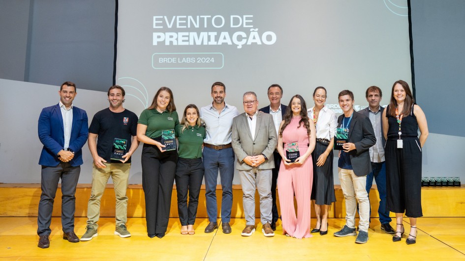 Foto posada de frente. Todos estão de pé, são cerca de 12 adultos. Leite está ao centro. Os vencedores carregam os prêmios. Todos sorriem.