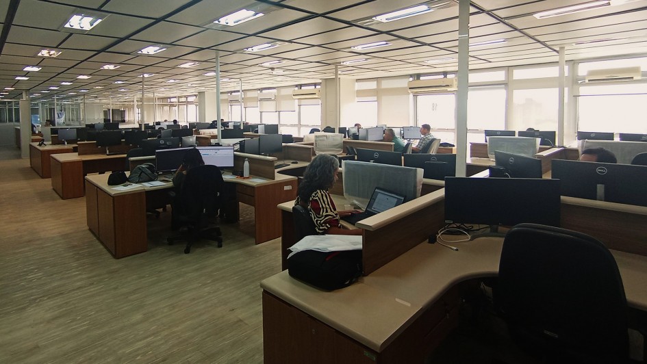 Imagem de uma ampla sala com estações de trabalho e pessoas trabalhando nelas.