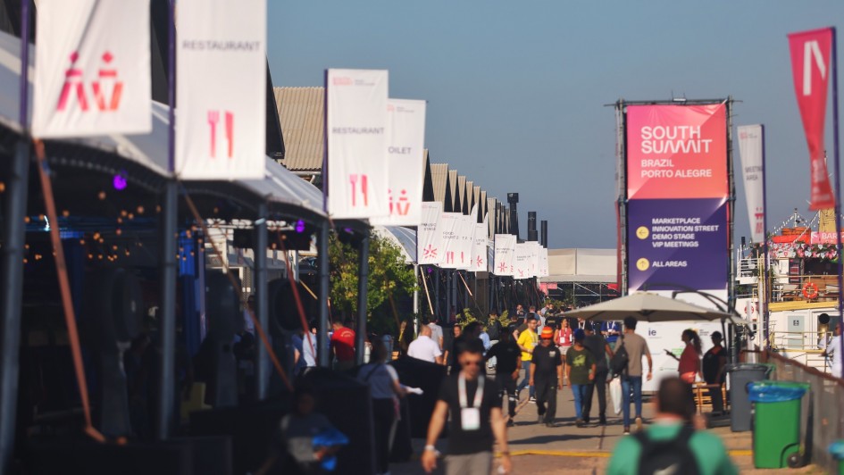 A imagem mostra uma área externa do evento South Summit Brazil, realizado em Porto Alegre. Há diversas pessoas circulando pelo espaço, que está decorado com placas e banners informativos. No centro, um grande banner indica as direções para áreas como Marketplace, Innovation Street, Demo Stage e VIP Meetings. Tendas com sinalização de restaurante, banheiro e palcos estão alinhadas à esquerda.