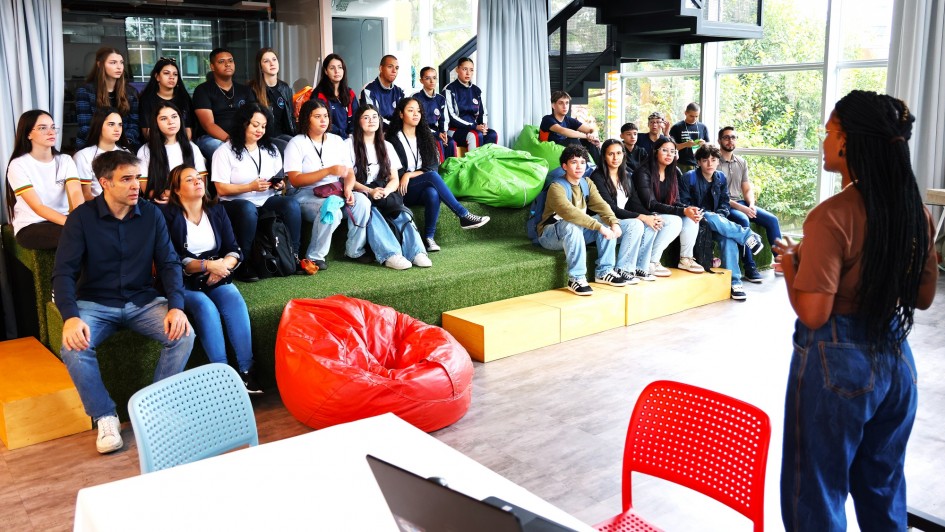 Foto de jovens participantes sentados em um tablado assitindo a uma palestra no HackaTchê Business 2025. A palestrante pode ser vista de costas mais à direita da imagem.