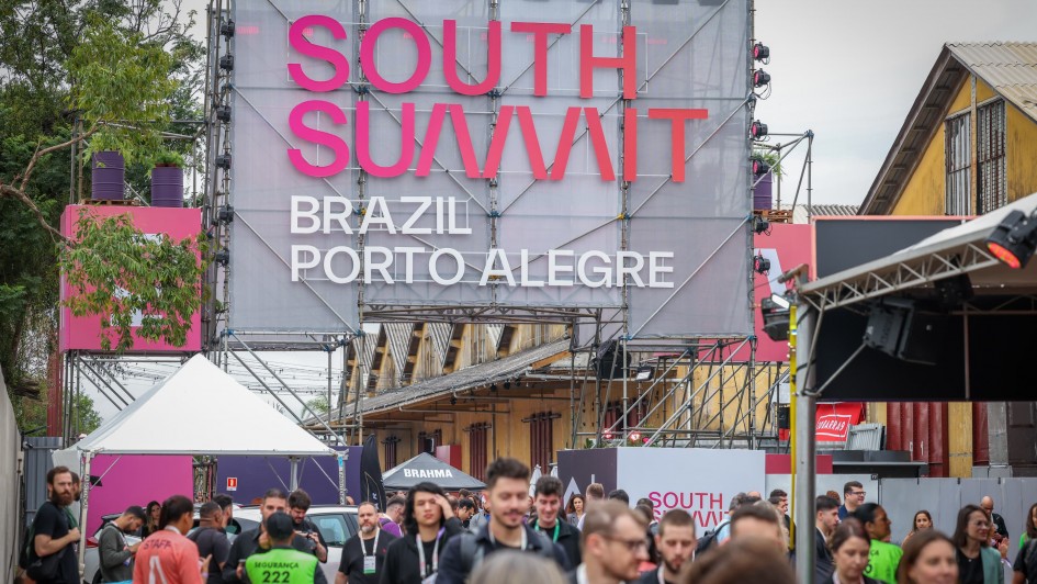 A imagem mostra a entrada do South Summit Brazil 2025. O portal do evento é montado com uma estrutura metálica moderna e decorado com plantas em vasos coloridos, elementos visuais vibrantes e painéis com logotipos. Ao fundo, é possível ver pessoas caminhando pelo espaço do evento.